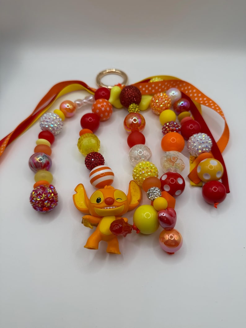 May include: A handmade keychain featuring an orange Stitch figurine and a variety of colorful beads in shades of red, orange, and yellow. The keychain includes a gold-colored ring and orange ribbons with polka dots.