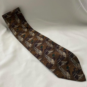 Puede incluir: Corbata marrón con un patrón repetido de patos en vuelo. La corbata tiene un fondo marrón oscuro con patos en tonos marrones, blancos y verdes. La corbata está en ángulo sobre una superficie blanca.