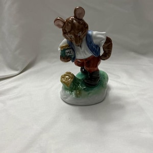 May include: A ceramic figurine of a bear wearing a white shirt, blue vest, and red pants, holding a bag labeled "MEZ". The bear stands on a green and white base, with a brown body and ears.