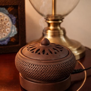 May include: A brown ceramic incense burner with a perforated body and a decorative lid. The burner is emitting a wisp of smoke, suggesting it is in use. The design includes a base and a lid with cut-out patterns.