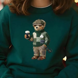 Könnte beinhalten: Ein dunkelgrünes Sweatshirt mit einem Cartoon-Bären, der einen Patchwork-Pullover, eine Mütze trägt und einen Becher hält. Der Bär hat ein Kleeblatt auf seinem Pullover und eine Brille, die an seiner Tasche hängt. Das Sweatshirt hat einen weißen Kragen.