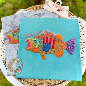 May include: Embroidered fish design on a teal fabric. The fish is a patchwork of orange, red, and blue, with the text "Catch ya later". A similar design is on a gray fabric. A collection of bracelets is in the lower left corner.