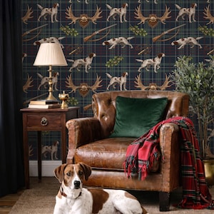 May include: A living room scene featuring a brown leather armchair with a green velvet pillow and a red and green plaid blanket. The background is a wallpaper with a hunting theme, showcasing dogs, antlers, and rifles. A dog is lying on the floor.