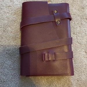 May include: A burgundy leather journal with a wraparound strap closure. The journal is rectangular and features a textured surface. The strap is secured with two brass-colored rivets and a buckle closure. The journal is closed.