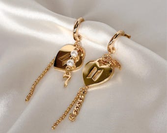 May include: A pair of gold-coloured heart-shaped earrings. Each earring features a heart pendant with the number 7 or 11, adorned with small pearls and dangling chain accents. The earrings are designed with a hoop closure.