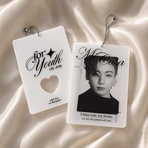 Könnte beinhalten: Zwei rechteckige Schlüsselanhänger aus weißem Acryl mit silbernen Kugelkettchen. Einer hat einen herzförmigen Ausschnitt und den Text "for Youth est. 2013 with luv, to Bangtan". Der andere zeigt ein Schwarz-Weiß-Porträt mit dem Text "Mi Casa" und "I miss you, my home. we are not seven with you".
