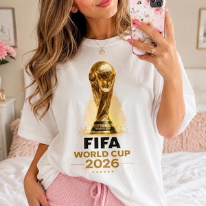 May include: White t-shirt featuring a golden FIFA World Cup trophy graphic. Below the trophy, the text reads "FIFA WORLD CUP 2026" in bold, black letters. The design includes a gold star pattern. A casual, sporty tee.