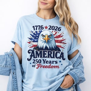 May include: Light blue t-shirt featuring a detailed illustration of an American bald eagle with the text "1776-2026 AMERICA 250 Years of Freedom". The design incorporates the American flag and stars.