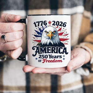 May include: White ceramic mug with a black handle and rim. The mug features a patriotic design with an eagle, the text "1776 2026 AMERICA 250 Years of Freedom" in red, white, and blue.