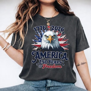 May include: Dark gray t-shirt featuring a patriotic design. The graphic includes a bald eagle, the American flag, and the text "1776 2026 AMERICA 250 YEARS OF FREEDOM". The eagle has white feathers and a yellow beak.