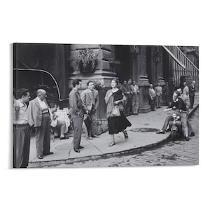 Op de afbeelding: Zwart-witfoto van een straatbeeld met verschillende mensen. Een vrouw in een donkere jurk loopt op de stoep. Mannen in pakken en vrijetijdskleding staan en praten. Een man rijdt op een scooter.