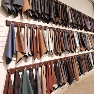 May include: A large wall display featuring three horizontal wooden racks holding numerous leather swatches. The swatches are arranged in rows, showcasing a wide variety of colors including shades of brown, gray, blue, black, and cream.