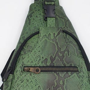 Może przedstawiać: Zielona torba typu sling z wzorem skóry węża, czarnym zamkiem błyskawicznym i zapięciem na klamrę. Torba ma pojedynczy regulowany pasek i przednią kieszeń zapinaną na zamek. Torba jest wykonana z wytrzymałego materiału i jest przeznaczona do noszenia na ciele.