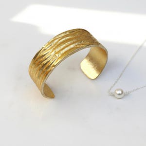 May include: A gold-coloured cuff bracelet with a textured, ridged surface sits beside a delicate silver necklace with a single white pearl pendant. The jewellery is displayed on a white surface.