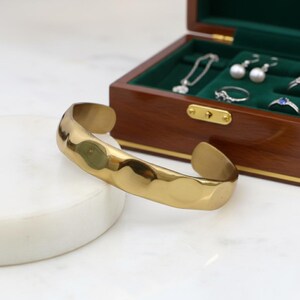 Puede incluir: Un brazalete dorado con textura martillada. El brazalete abierto se muestra sobre una superficie blanca, con una caja de joyería de madera en el fondo. La caja de joyería contiene otras joyas.