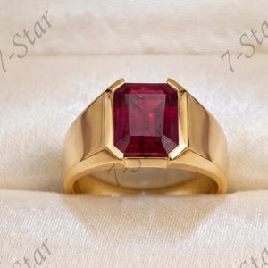 Op de afbeelding: Een gouden ring met een grote, rechthoekige, smaragdgeslepen dieprode edelsteen in een gladde, gepolijste gouden band. De ring wordt gepresenteerd in een donkerhouten juwelendoos met een crèmekleurige voering en de tekst 7-Star is zichtbaar.