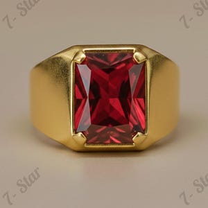 May include: A gold-colored ring with a large, rectangular, deep red gemstone. The ring has a classic, bold design with a polished finish. The gemstone is set in a bezel setting, enhancing its brilliance. The ring is a statement piece of jewelry.