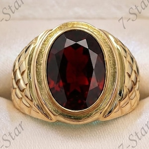 Puede incluir: Un anillo de oro con una gran piedra preciosa granate de color rojo intenso y corte ovalado, montado en una banda texturizada con patrón de tejido de cesta. El anillo se presenta en un joyero de madera oscura con forro crema y el texto 7-Star.