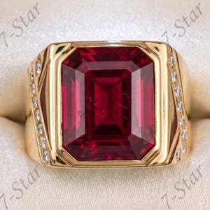 Könnte beinhalten: Ein Goldring mit einem großen, rechteckigen, tiefroten Edelstein in der Mitte. Das Band ist mit kleinen, klaren, runden Steinen besetzt, die auf beiden Seiten der zentralen Fassung angebracht sind. Der Ring wird in einer dunklen Holzschatulle präsentiert.