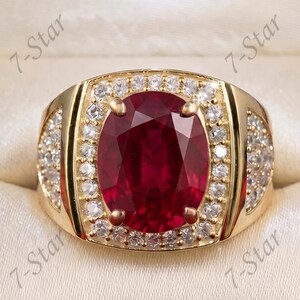 Könnte beinhalten: Ein Goldring mit einem großen, oval geschliffenen, tiefroten Edelstein in der Mitte, umgeben von einem Kranz aus kleinen, runden, klaren Steinen. Die Ringschiene ist breit und ebenfalls mit kleinen, klaren Steinen besetzt. Der Text 7-Star ist sichtbar.