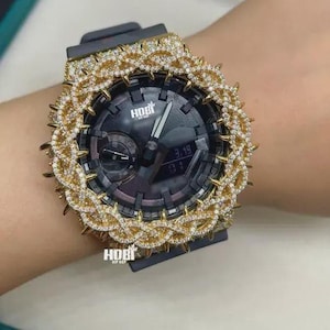 May include: A black analog-digital wristwatch featuring a prominent gold-tone bezel encrusted with numerous small clear stones in a twisted, spiked pattern. The watch face displays the text HOBI and includes both hands and a digital window.