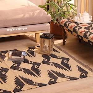 May include: A rectangular area rug featuring a repeating black geometric pattern on a natural beige background. The rug has a textured weave with fringed edges. A small stack of books rests on the surface, and a wooden lantern sits nearby.
