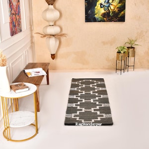 May include: A rectangular area rug featuring a dark grey base with a repeating white geometric pattern. The rug is positioned on a white floor and includes the text RugStoreJaipur printed in the bottom right corner of the rug surface.