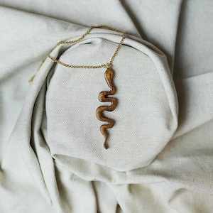 Op de afbeelding: Halsketting met houten slangenhanger en goudkleurige ketting. De hanger is in slangen vorm gesneden en toont de natuurlijke houtnerf. De ketting ligt op een neutraal gekleurde stof, wat een eenvoudige, elegante presentatie creëert.