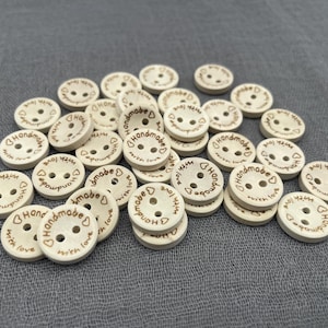 Pode incluir: Uma coleção de botões de madeira de cor clara com dois furos. Cada botão é gravado com as palavras "Handmade with love" em torno de um símbolo de coração. Os botões estão espalhados sobre um tecido cinzento.
