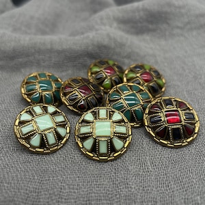 May include: A collection of decorative buttons with a gold-tone border. The buttons feature a mosaic design in shades of green, red, black, and light green. The buttons are arranged on a gray fabric surface.