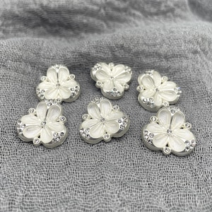 May include: Six white flower-shaped buttons with silver-colored edges and clear crystal accents. The buttons have a three-dimensional design and are arranged on a gray fabric surface. These decorative buttons are ideal for sewing and craft projects.