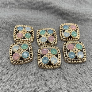 May include: Six square decorative buttons with a gold-coloured frame and a pattern of four colourful, iridescent gems. The gems are in shades of pink, blue, and green. The buttons are on a grey fabric surface.
