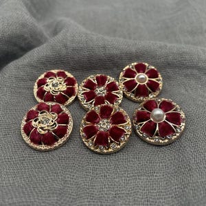 May include: Six decorative buttons with a floral design. The buttons have a gold-colored rim, a deep red velvet-like center, and are adorned with rhinestones and a pearl. The buttons are arranged on a gray fabric.