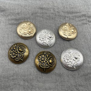 Op de afbeelding: Zes ronde knopen met een verhoogd bloemmotief van een roos en een madeliefje. De set bevat twee matgouden, twee zilveren en twee donkerbronzen knopen met een textuurafwerking, die allemaal hetzelfde in reliëf gemaakte bloemmotief vertonen.