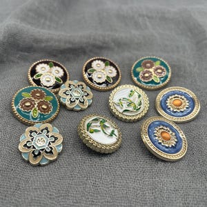 May include: A selection of decorative buttons with floral and botanical designs. The buttons feature a variety of colours, including white, black, teal, and gold. Some buttons are round, others oval.