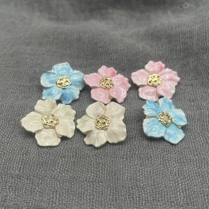 May include: Six flower-shaped decorative embellishments in shades of blue, pink, and cream, each with a gold-colored center. The flowers are arranged on a gray fabric surface, showcasing their delicate design.