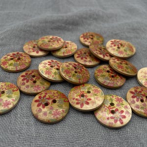 Pode incluir: Uma coleção de botões de madeira com um design floral. Os botões redondos, com dois ou quatro furos, apresentam flores vermelhas e rosa sobre um fundo bege claro. Estão dispostos sobre uma superfície de tecido cinzento.