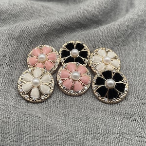 May include: A collection of six decorative flower-shaped buttons. The buttons feature a central pearl surrounded by petals in pink, black, and white. Each button has a gold-colored rim with small, clear rhinestones.