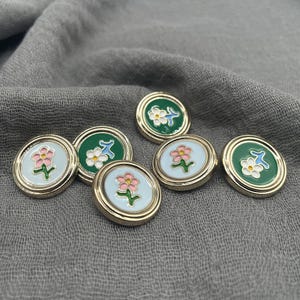 Op de afbeelding: Een verzameling decoratieve knopen met bloemmotieven. De knopen hebben een goudkleurige metalen rand en hebben een lichtblauwe of groene achtergrond met roze of witte bloemen. De knopen zijn gerangschikt op een grijze stof.