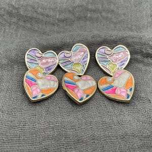 Puede incluir: Seis broches en forma de corazón con bordes dorados. Los broches presentan diseños de esmalte coloridos en tonos de naranja, rosa, azul, morado y amarillo. Los broches están dispuestos sobre una tela gris.