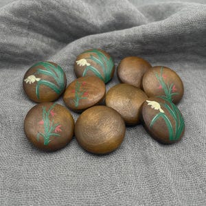 May include: A collection of nine round, brown decorative stones. Each stone features a hand-painted floral design in green, white, and red. The stones are arranged on a gray, textured surface.