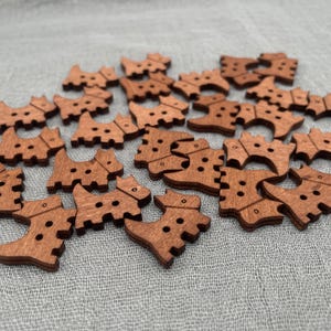 以下が含まれることがあります： 犬の形をした小さな木製ボタンのコレクション。ボタンは温かみのある茶色で、縫い付け用の穴が2つまたは3つあります。明るい灰色の布の上に散らばっています。