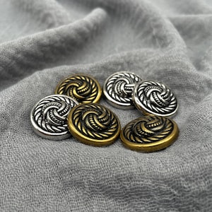 May include: A collection of six decorative buttons in silver and gold tones. Each button features a spiral design with a textured border. The buttons are arranged on a gray fabric background.