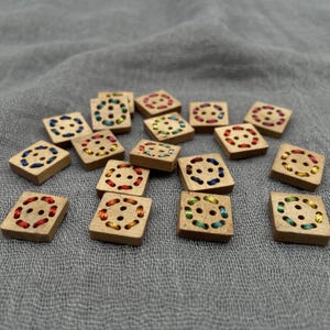 May include: A selection of square wooden buttons with colourful thread detailing. Each button has a stitched design around the buttonholes, with threads in various colours such as red, blue, yellow, and green. The buttons are arranged on a grey fabric surface.