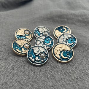 May include: A collection of round, decorative buttons with a celestial theme. Each button features a teal enamel border, a crescent moon, and a pearl-like center. The buttons have gold and silver metallic finishes.