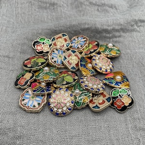 May include: A selection of decorative buttons with detailed designs. The buttons display floral, fruit, and geometric patterns in colours such as red, pink, blue, and green, with gold-coloured metal rims. Some buttons have pearl accents.
