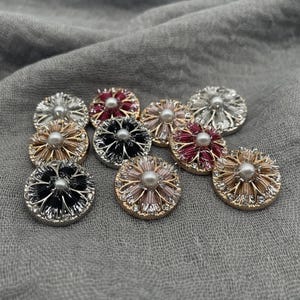 Puede incluir: Un conjunto de botones decorativos con forma de flor en plata, dorado, negro y burdeos. Cada botón presenta una perla central y un borde con pedrería. Ideal para adornar prendas o manualidades.