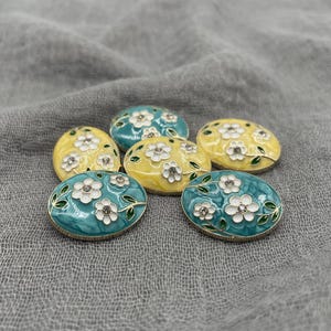 May include: Six oval-shaped decorative buttons. The buttons feature a floral design with white flowers and green leaves on a yellow or teal background. The buttons have a gold-colored trim.