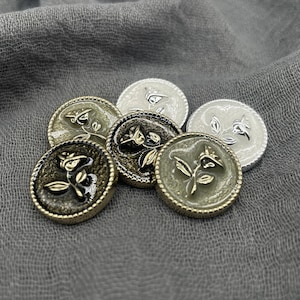 May include: Six decorative buttons with a floral design and beaded edges. The buttons are in silver, gold, and olive green metallic finishes. They are arranged on a grey fabric. Each button has a diameter of approximately 2.5 cm.