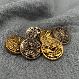 May include: A selection of round, decorative buttons in gold and silver hues. Each button showcases a detailed relief of a classical figure with laurel leaves. The buttons are displayed on a textured, grey fabric.